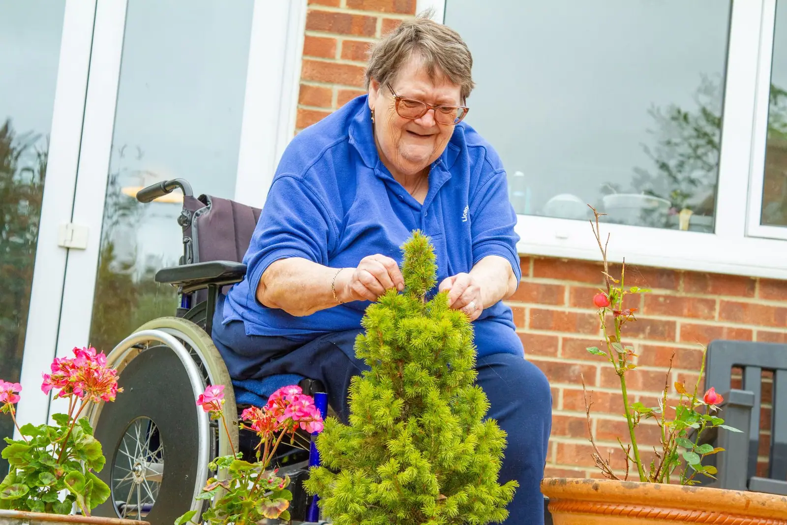 Smiling older woman in a wheelchair tending potted plants outside a brick house (representative photo)